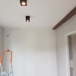 ALL-BUD Arkadiusz Bednarek - Wnętrze pokoju po remoncie z szarą ścianą, dwiema czarnymi lampami sufitowymi, dekoracyjną brązową belką pod sufitem oraz drabiną malarską w rogu.