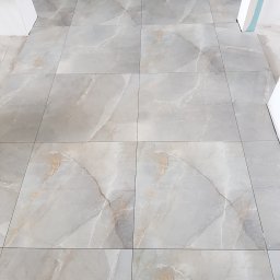 ALL-BUD Arkadiusz Bednarek - Ułożone szare płytki podłogowe imitujące marmur z widocznymi fugami, fragment progu drzwiowego w tle, pozostałości po pracach wykończeniowych.