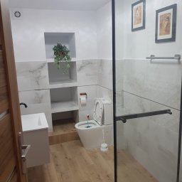 ALL-BUD Arkadiusz Bednarek - Jasna łazienka po remoncie: drewnopodobna podłoga, białe płytki imitujące marmur, kabina prysznicowa ze szkła, umywalka z szafką i WC w zabudowie.