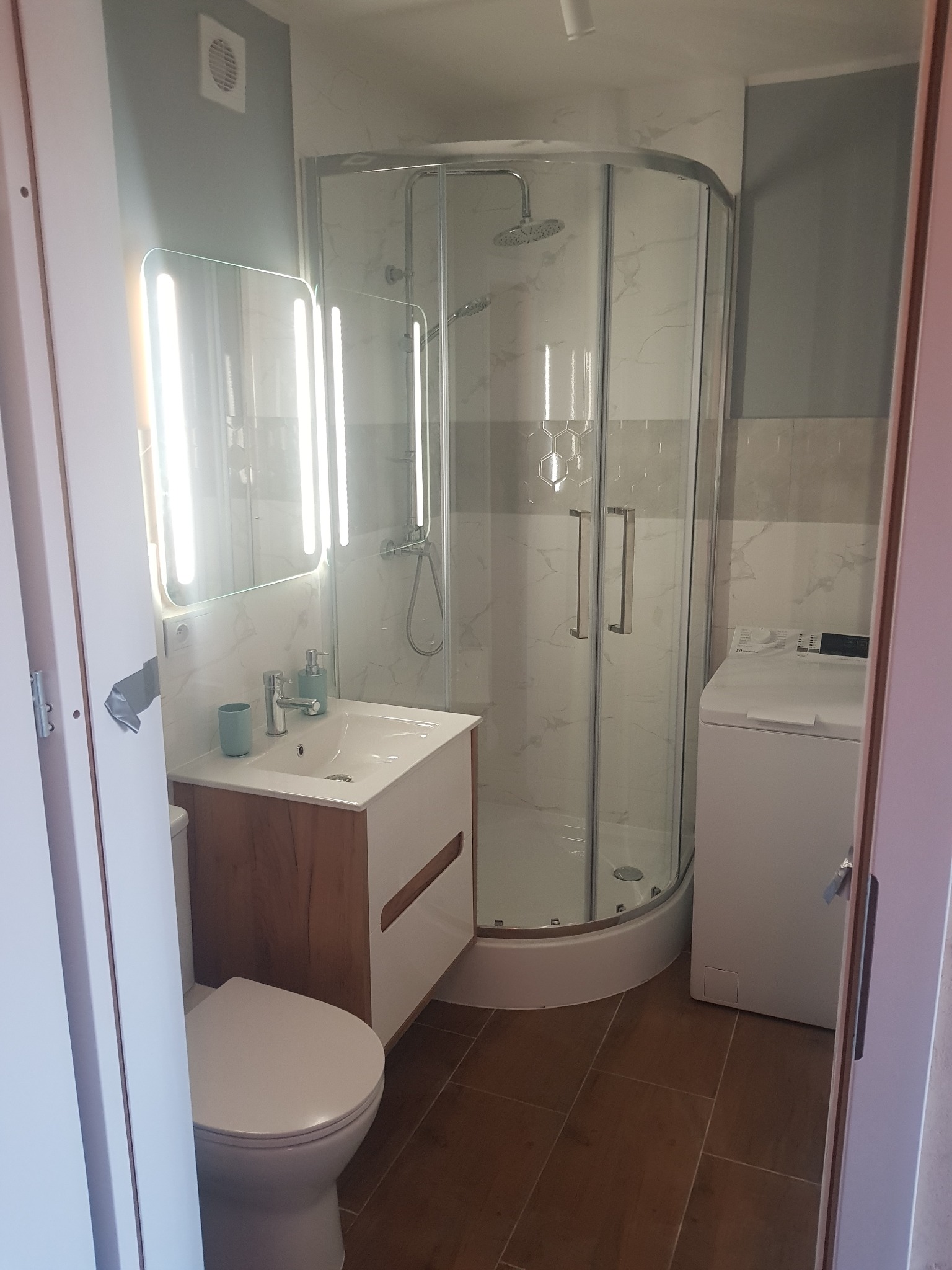 Nowoczesna łazienka po remoncie: kabina prysznicowa z hydromasażem, umywalka z szafką z drewnopodobnym frontem, podświetlane lustro, pralka i brązowe płytki podłogowe.