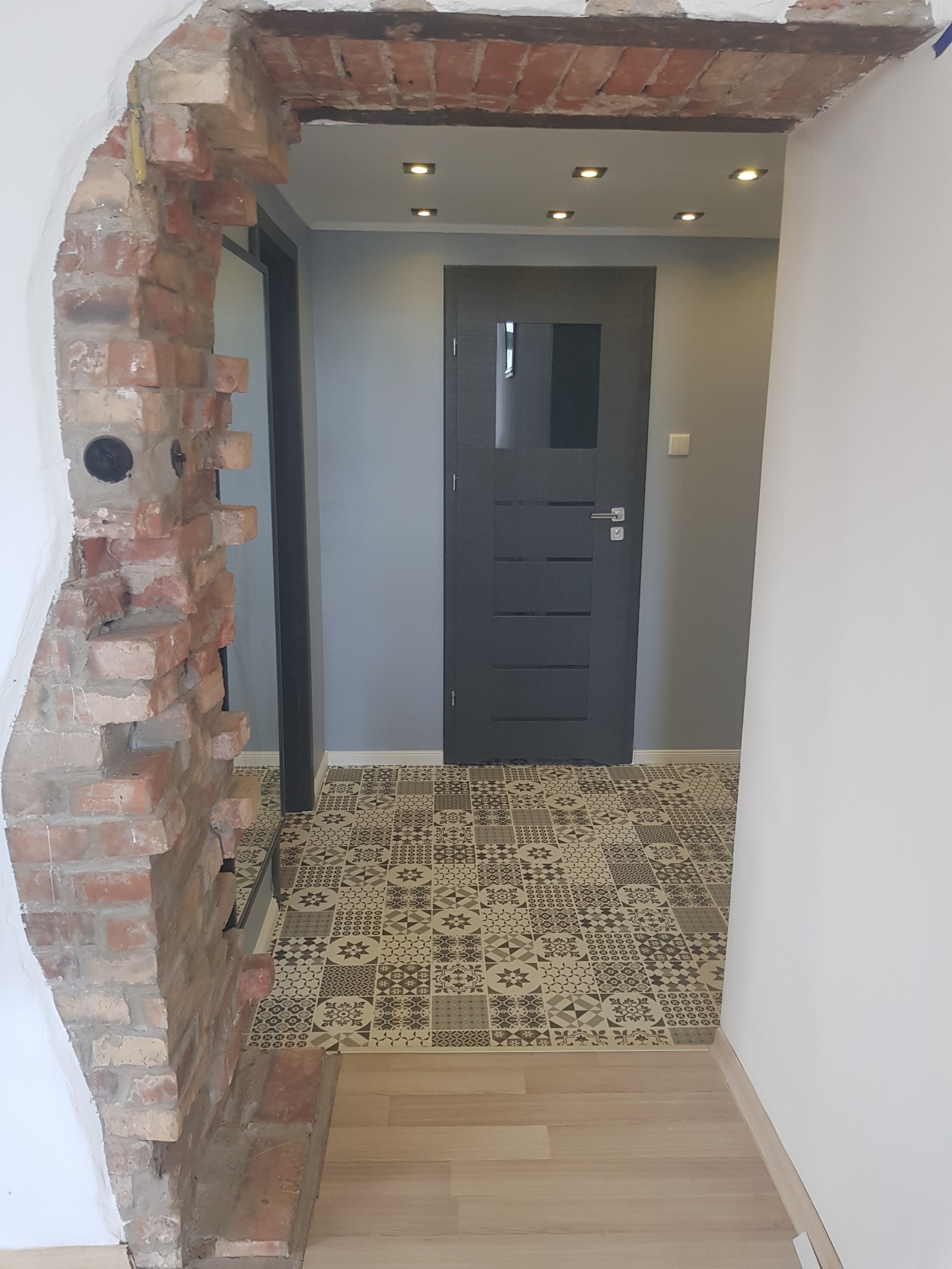 Otwór w ścianie z odsłoniętą cegłą w Suwałkach, prowadzący do przedpokoju z szarą ścianą, wzorzystą podłogą i ciemnymi drzwiami.