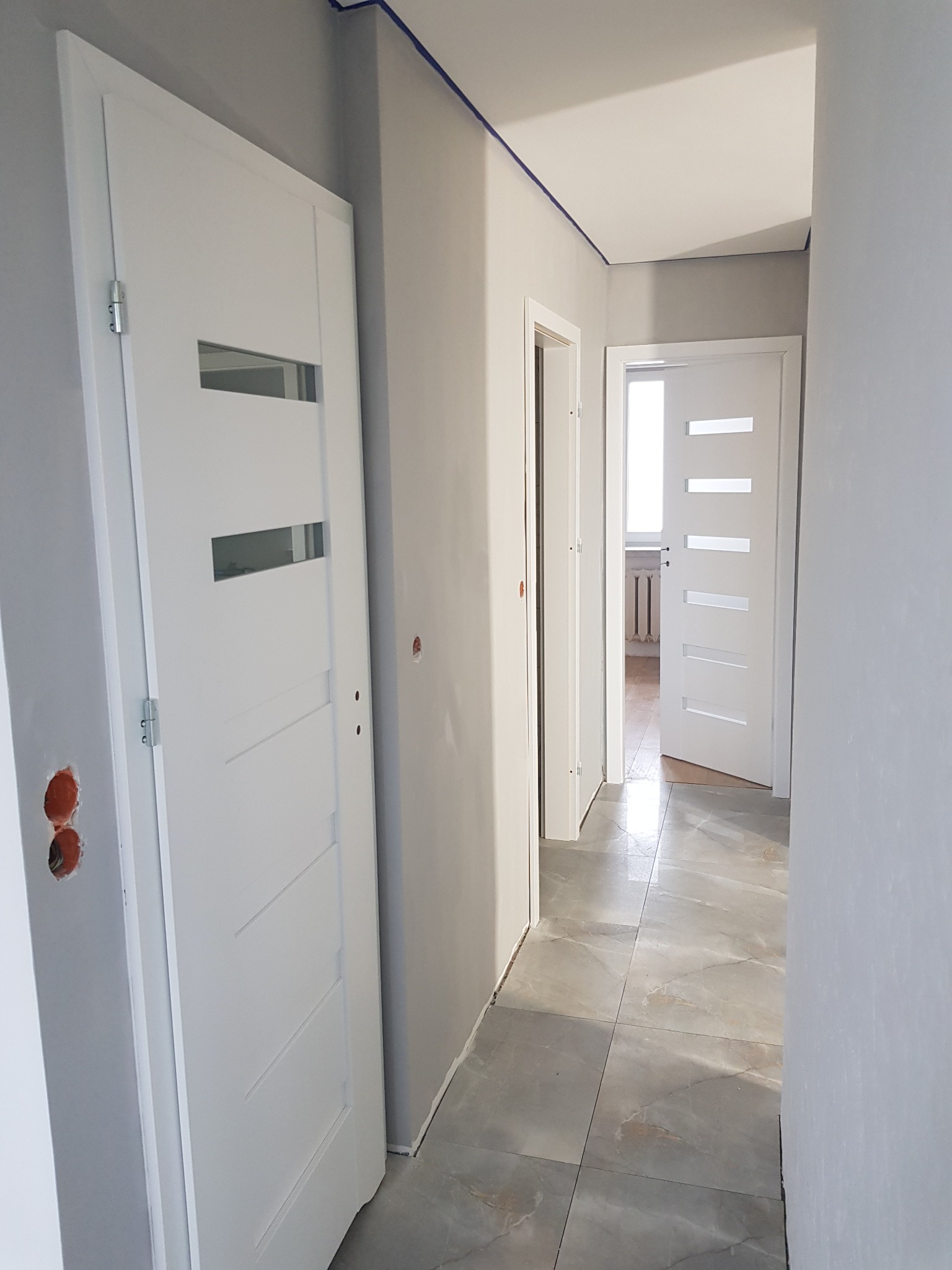 Korytarz w trakcie prac wykończeniowych: szare ściany z niezakrytymi otworami na gniazdka, ułożone szare płytki podłogowe imitujące marmur, białe drzwi wewnętrzne z przeszkleniami, zabezpieczenia...