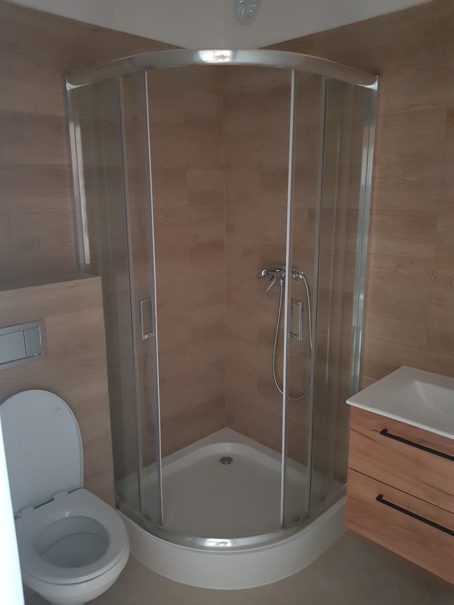 Nowoczesna łazienka z kabiną prysznicową narożną z chromowanymi profilami, podwieszaną toaletą i szafką pod umywalkę z drewnopodobnym frontem.