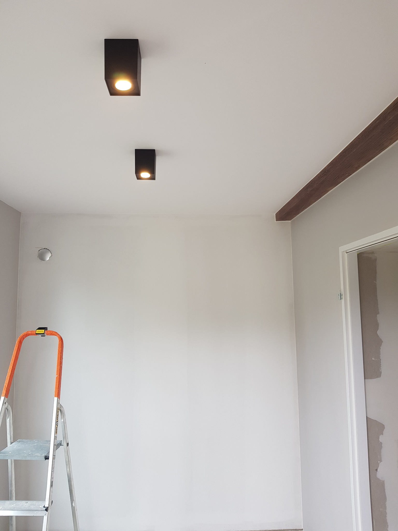 Wnętrze pokoju po remoncie z szarą ścianą, dwiema czarnymi lampami sufitowymi, dekoracyjną brązową belką pod sufitem oraz drabiną malarską w rogu.