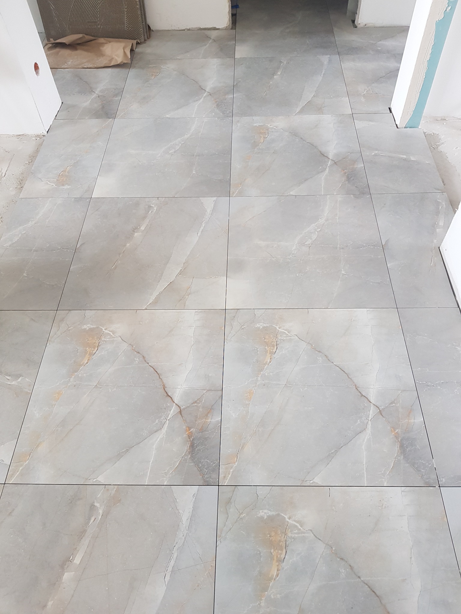 Ułożone szare płytki podłogowe imitujące marmur z widocznymi fugami, fragment progu drzwiowego w tle, pozostałości po pracach wykończeniowych.