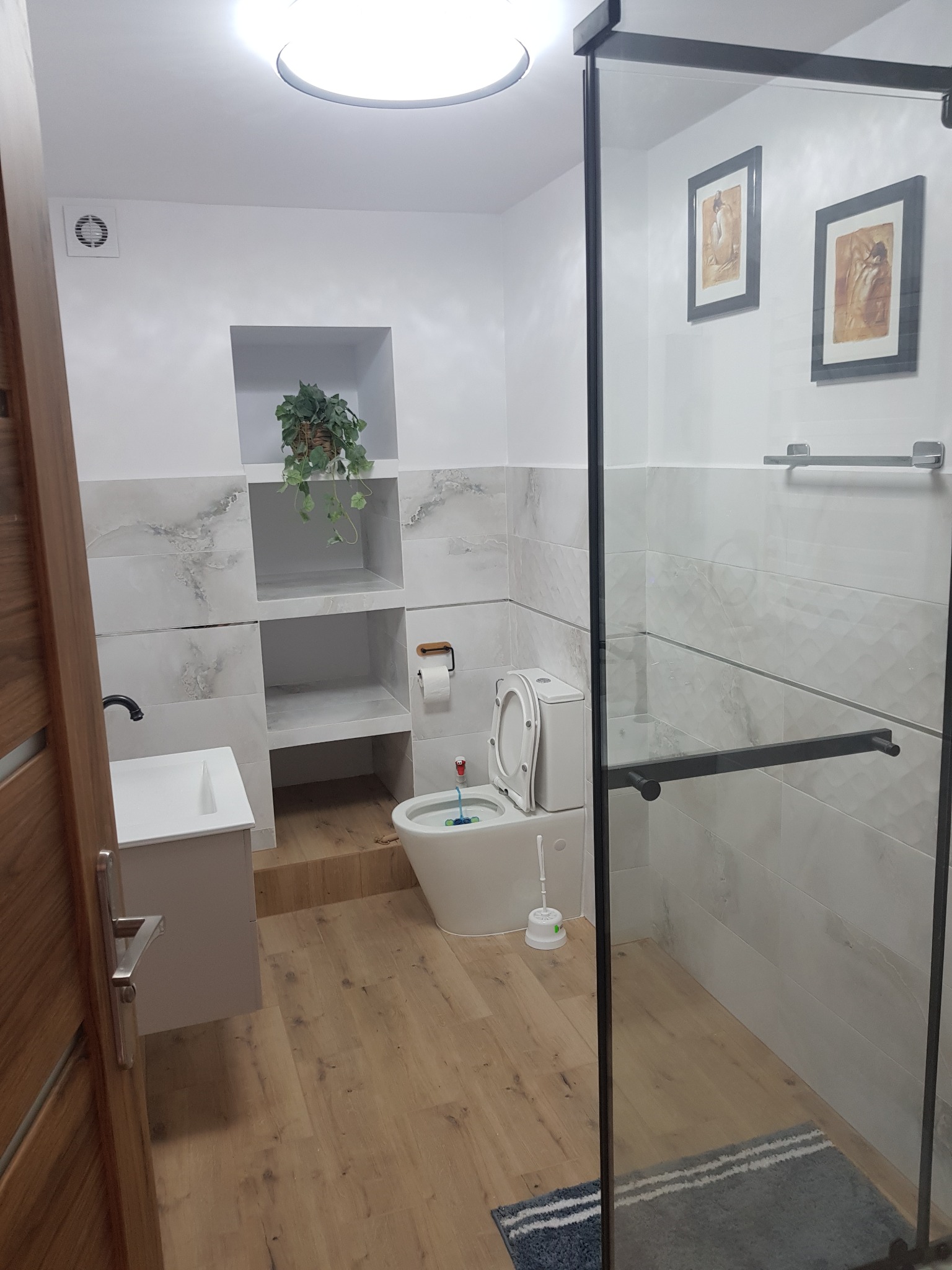 Jasna łazienka po remoncie: drewniana podłoga, białe płytki imitujące marmur, kabina prysznicowa z czarnymi ramami, umywalka i toaleta w minimalistycznym stylu.