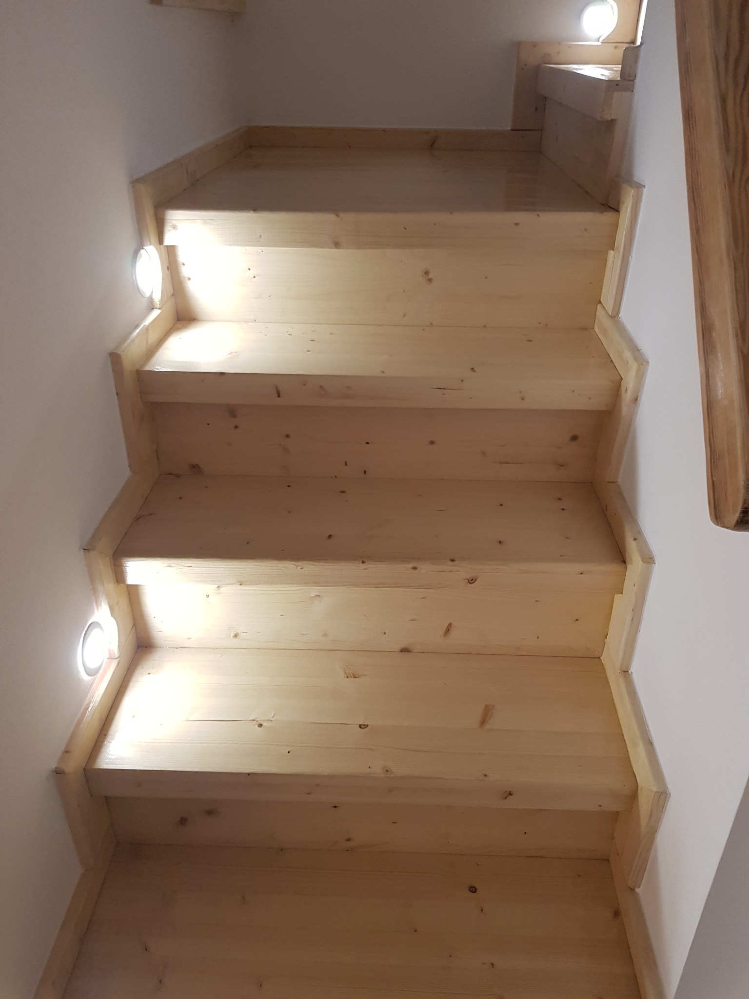 Jasne, drewniane schody z oświetleniem punktowym wzdłuż ściany, widok od dołu. Naturalne wykończenie drewna, minimalistyczny design.