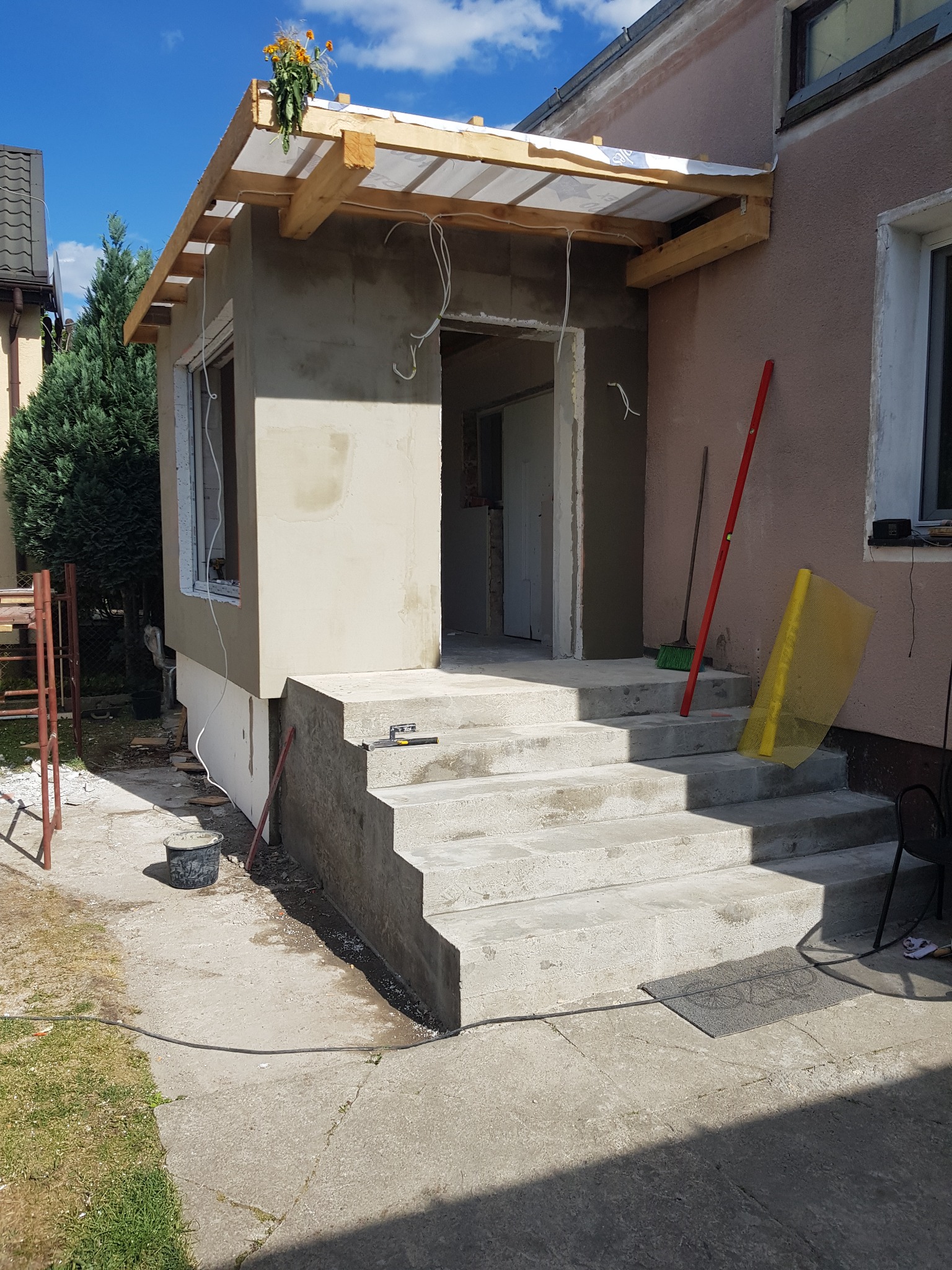 Budowa ganku z betonowymi schodami i drewnianą konstrukcją zadaszenia, widoczne narzędzia i materiały budowlane. Ściany w trakcie prac wykończeniowych.
