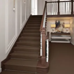 Klasyczna klatka schodowa z brązową wykładziną dywanową, białe ściany z dekoracyjnymi listwami i eleganckie oświetlenie ścienne. Widok na półpiętro z balustradą.