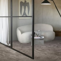 FloorArt - Wnętrze z miękką, szarą wykładziną dywanową w Kołobrzegu, oddzielone szklaną ścianą. Widoczny fotel, stolik i lampa, tworzące przytulną aranżację.