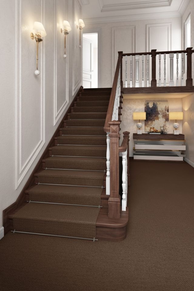 Klasyczna klatka schodowa z brązową wykładziną dywanową, białe ściany z dekoracyjnymi listwami i eleganckie oświetlenie ścienne. Widok na półpiętro z balustradą.