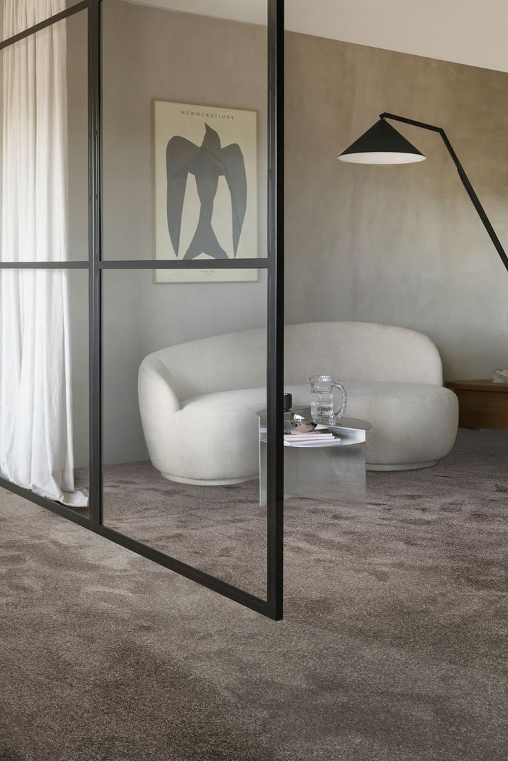 Wnętrze z miękką, szarą wykładziną dywanową w Kołobrzegu, oddzielone szklaną ścianą. Widoczny fotel, stolik i lampa, tworzące przytulną aranżację.