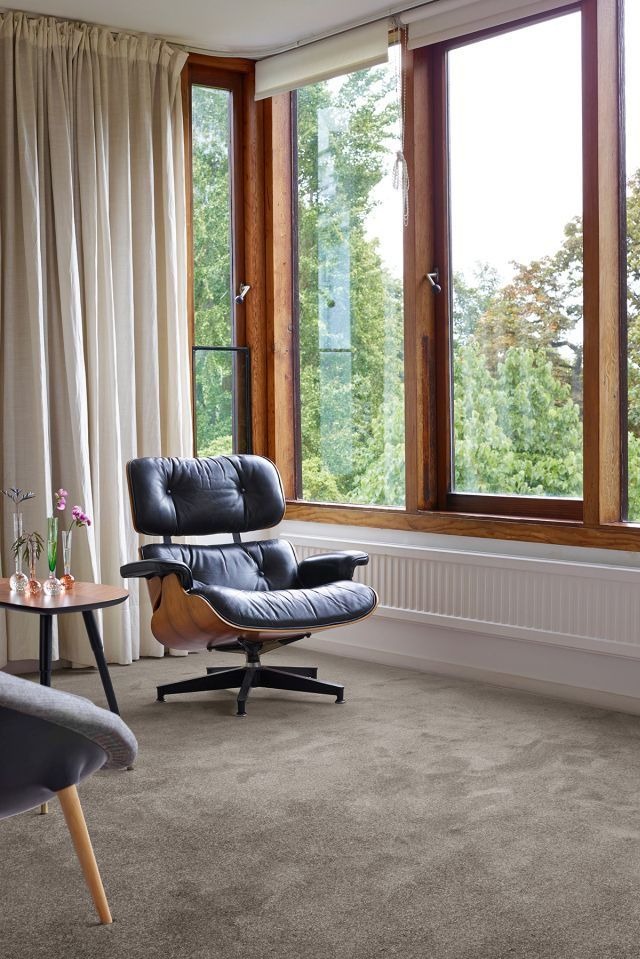 Stylowe wnętrze z szarą wykładziną dywanową, czarnym fotelem Eames i dużym oknem z drewnianą ramą. W tle zieleń drzew za oknem, nowoczesny grzejnik i stolik z wazonami.