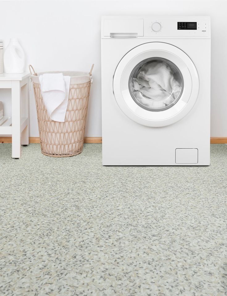 Jasna, wzorzysta wykładzina w pralni z pralką, koszem na pranie i stolikiem. Neutralna kolorystyka. Widok na całą podłogę, bez elementów montażowych.