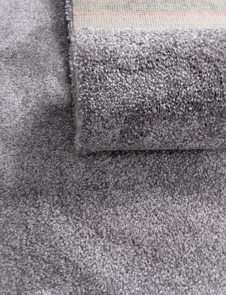 Szary dywan z krótkim włosiem, częściowo zwinięty, ukazujący strukturę podłoża. Delikatna faktura i stonowana kolorystyka.