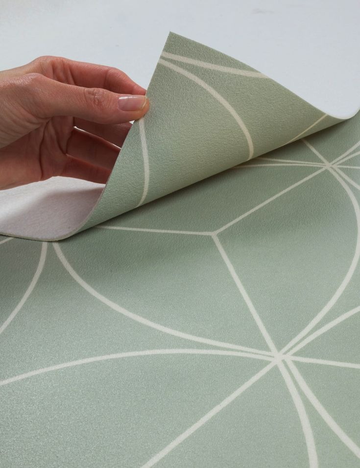 Wzorzysta wykładzina w odcieniach zieleni z geometrycznym motywem, podnoszona przez dłoń, ukazująca spodnią warstwę. Prezentacja produktu.