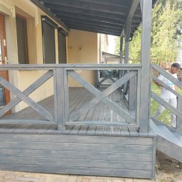 M-campentry - Szary taras drewniany z balustradą krzyżową, widok z boku, mężczyzna w tle wchodzi po schodach.