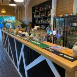 Drewniana konstrukcja baru z czarnymi panelami i białymi krzyżowymi wzmocnieniami, oświetlona wiszącymi lampami, z półkami pełnymi butelek w tle i dystrybutorami piwa na blacie.