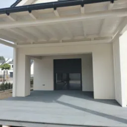 Biała konstrukcja z zadaszeniem, szary taras z desek, nowoczesny design, widok z zewnątrz, w tle domy jednorodzinne.