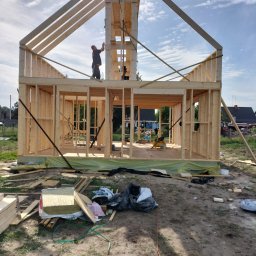 M-campentry - Wznoszenie surowej konstrukcji domu szkieletowego z drewna, widoczny pracownik na górnej belce, rozłożone materiały izolacyjne i odpady budowlane na ziemi wokół budynku.