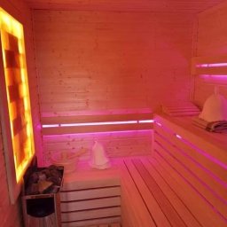 M-campentry - Wnętrze sauny wykończone jasnym drewnem, oświetlone nastrojowym, pomarańczowo-różowym światłem. Widoczne drewniane ławki, piec z kamieniami, drewniane wiadro z chochlą oraz filcowy kapelusz na głowę.
