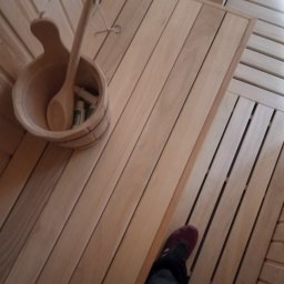 M-campentry - Wnętrze sauny z drewnianym wykończeniem, widoczny ceber z chochlą, butelki kosmetyków i fragment czyjejś nogi w ciemnych spodniach i bordowym bucie.