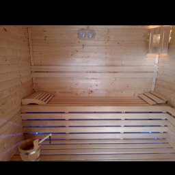 M-campentry - Wnętrze sauny wyłożone jasnym drewnem, z drewnianymi ławkami podświetlonymi na niebiesko, drewniany ceber z chochlą w rogu, dwa okrągłe termometry na ścianie.