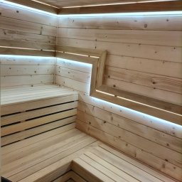 M-campentry - Wnętrze sauny wykończone jasnym drewnem, z podświetleniem LED wzdłuż górnej krawędzi i w narożnikach, ukazujące ławki i podłogę z desek.