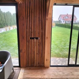M-campentry - Nowoczesna weranda z drewnianą podłogą i ścianą z pionowych desek, widok na zadbany trawnik i dom w tle, szare krzesło w rogu kadru.