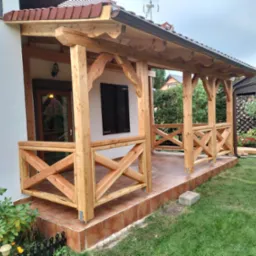 Zadaszony drewniany ganek z balustradą w kształcie krzyży, wykończony brązową dachówką, przylegający do białego budynku z brązowymi drzwiami i okiennicami, otoczony zielonym trawnikiem.