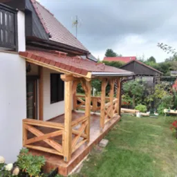 Zadaszony, drewniany taras przylegający do domu z białymi ścianami, widok z ogrodu, z elementami drewnianej konstrukcji w stylu rustykalnym i brązową terakotą na podłodze.