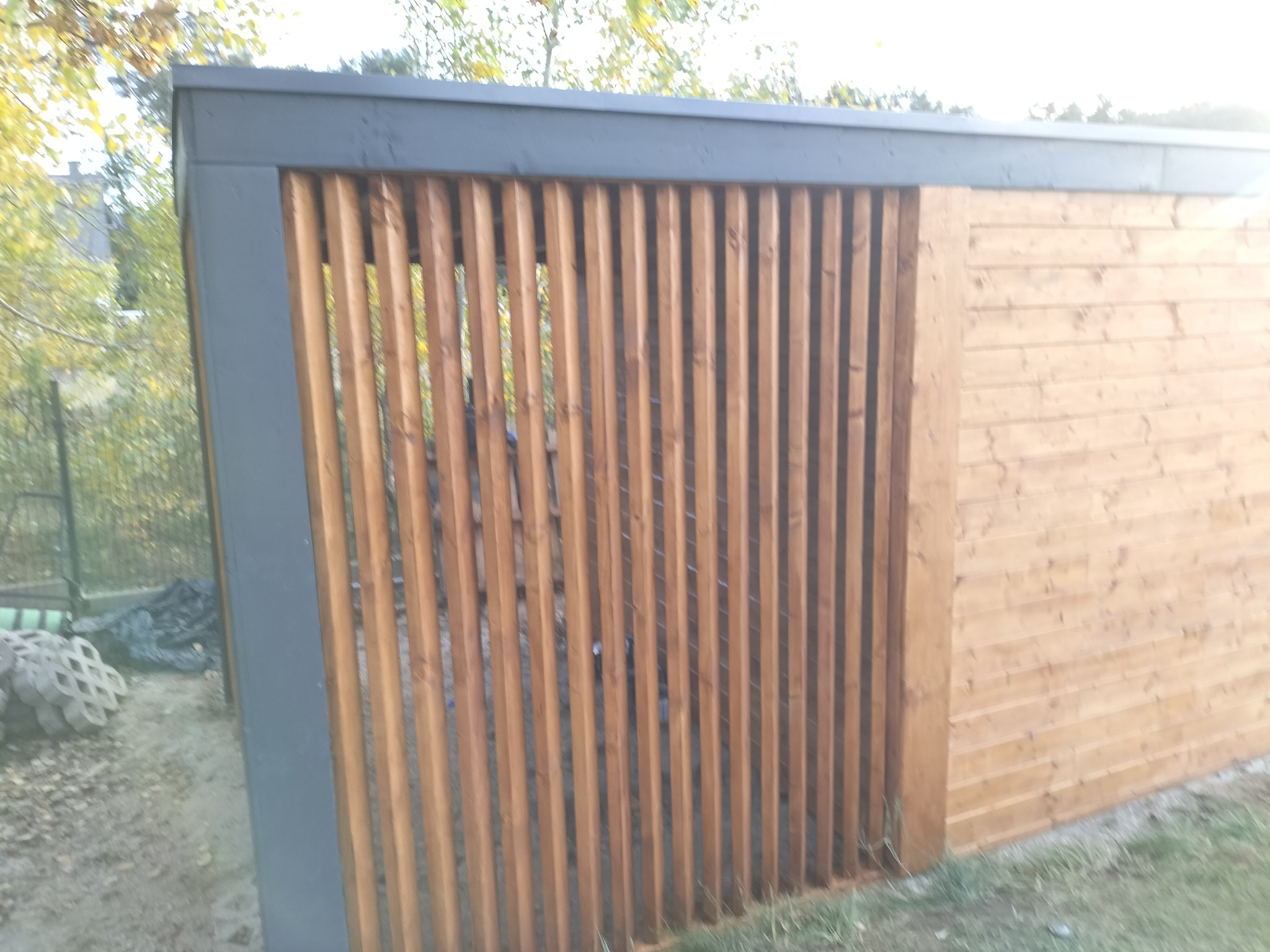 Nowoczesna konstrukcja z drewnianych listew i szarej ramy, tworząca ścianę lub element ogrodzenia, z widocznymi betonowymi elementami podłoża i drzewami w tle.