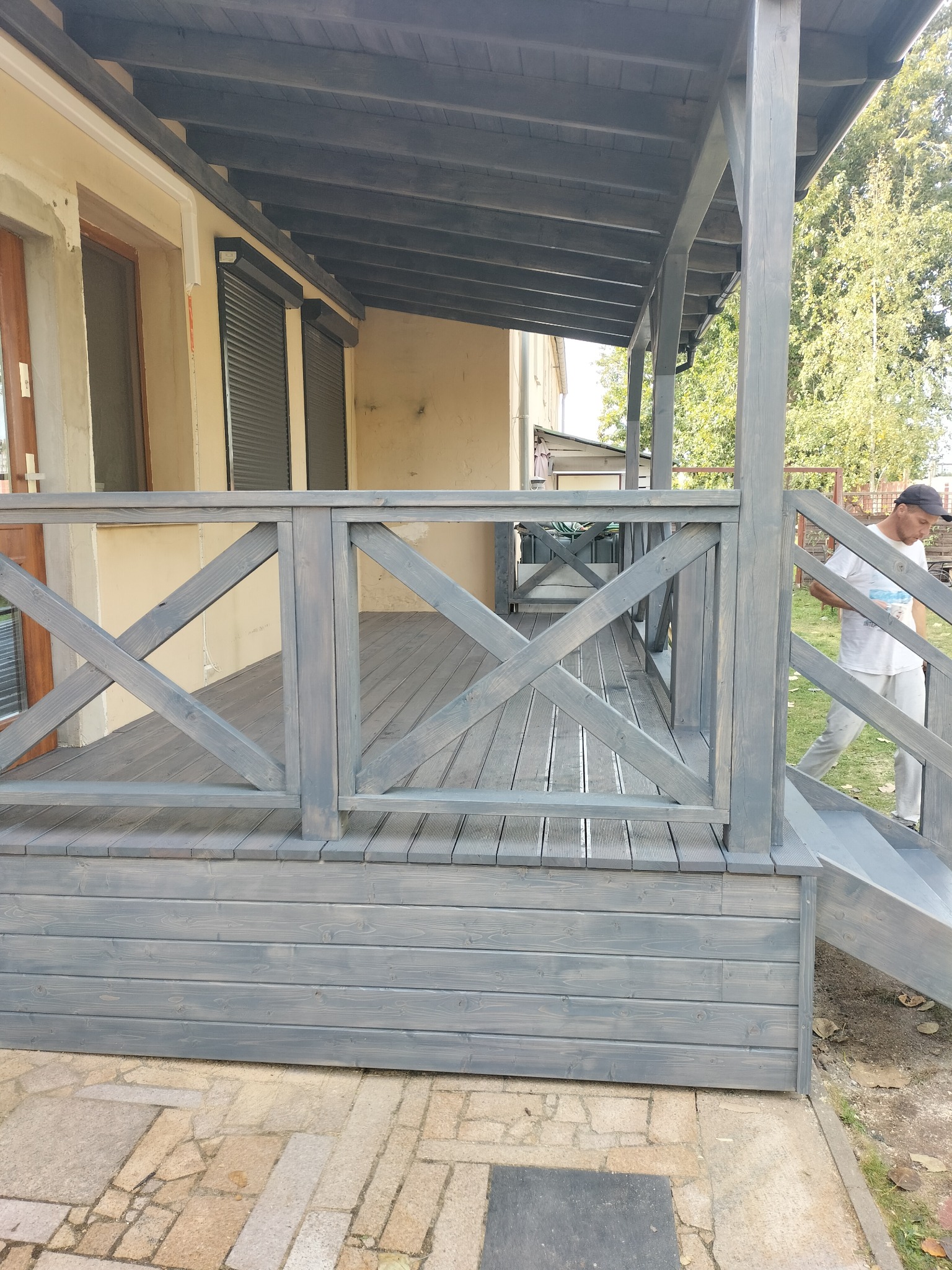 Szary taras drewniany z balustradą krzyżową, widok z boku, mężczyzna w tle wchodzi po schodach.