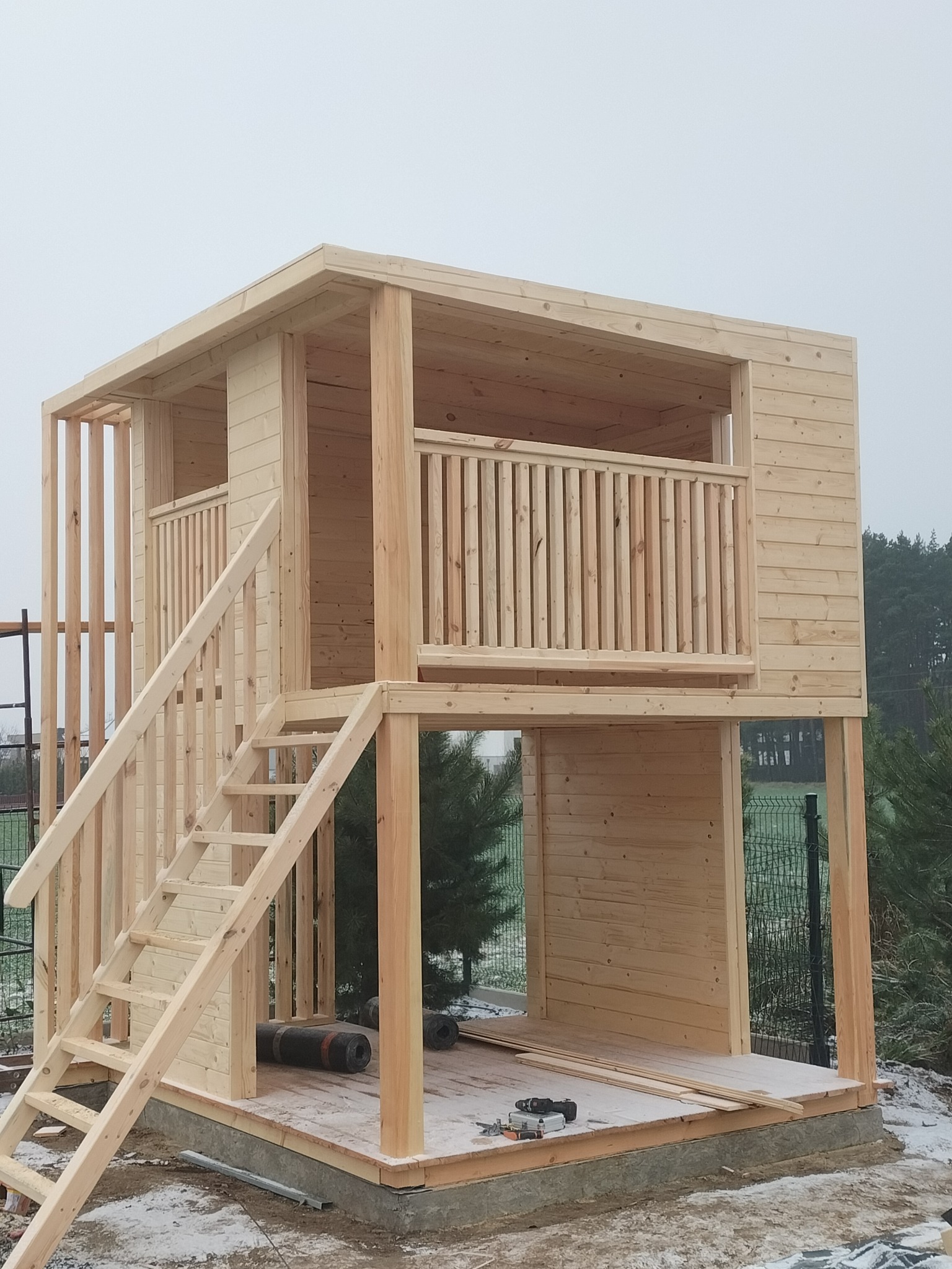 Surowa, dwupoziomowa konstrukcja z jasnego drewna w trakcie budowy, z widocznymi schodami, balustradą na górnym poziomie oraz narzędziami i materiałami budowlanymi na podłodze, otoczona zimowym...