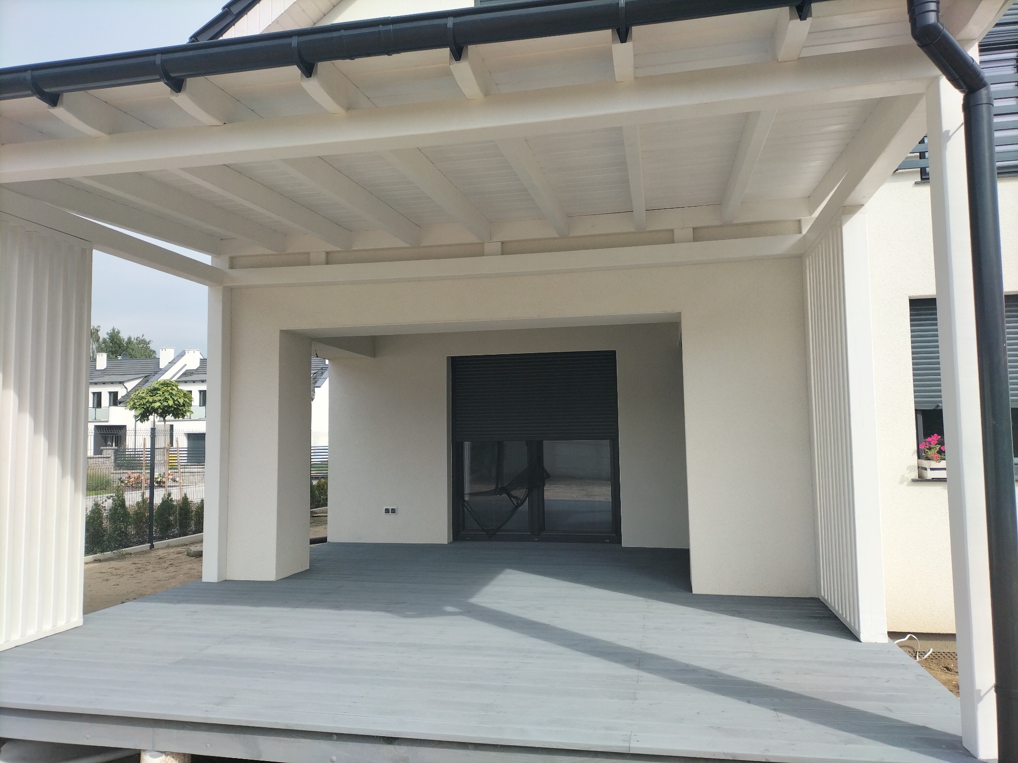 Biała konstrukcja z zadaszeniem, szary taras z desek, nowoczesny design, widok z zewnątrz, w tle domy jednorodzinne.