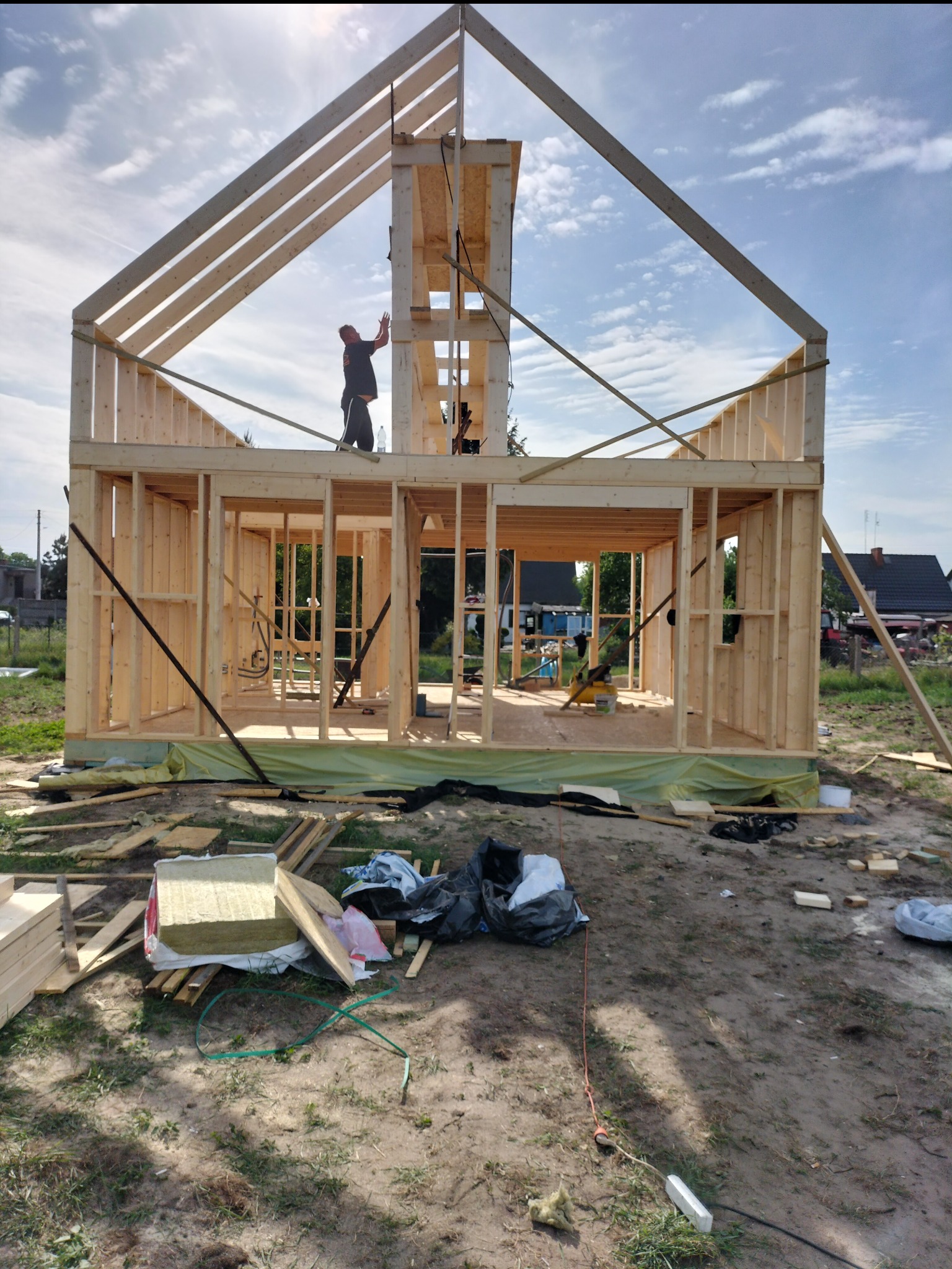 Wznoszenie surowej konstrukcji domu szkieletowego z drewna, widoczny pracownik na górnej belce, rozłożone materiały izolacyjne i odpady budowlane na ziemi wokół budynku.