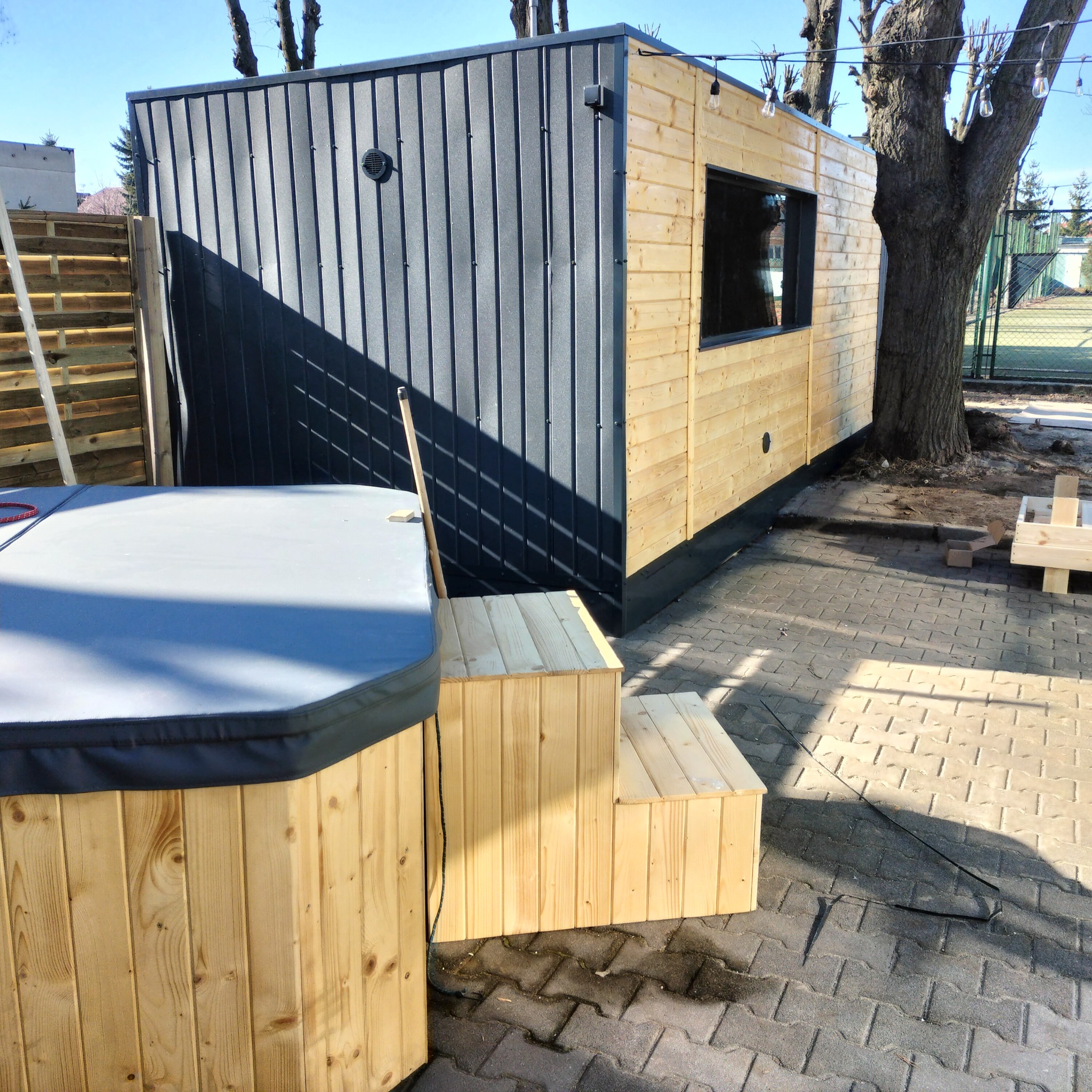 Nowoczesna, modułowa konstrukcja z drewna i blachy falistej, z oknem i schodami, obok jacuzzi z pokrywą, na tle kostki brukowej i drzewa z lampkami.