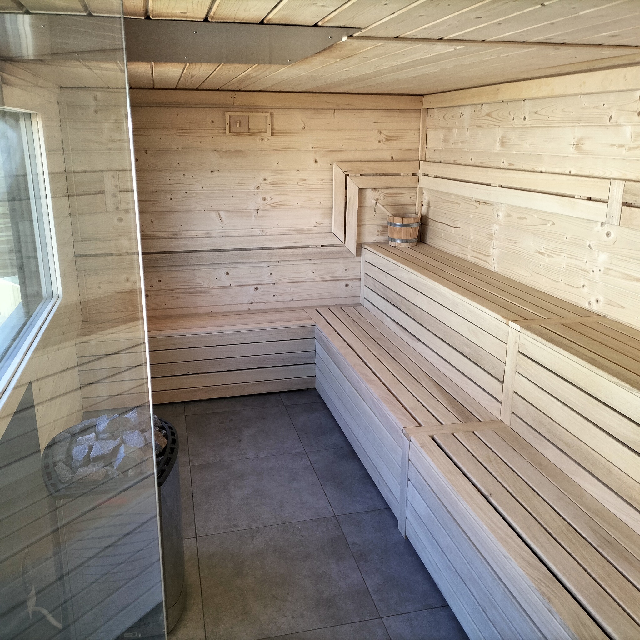 Wnętrze sauny z jasnego drewna z piecem na kamienie za szklaną ścianą, drewniany ceber wiszący na ścianie.