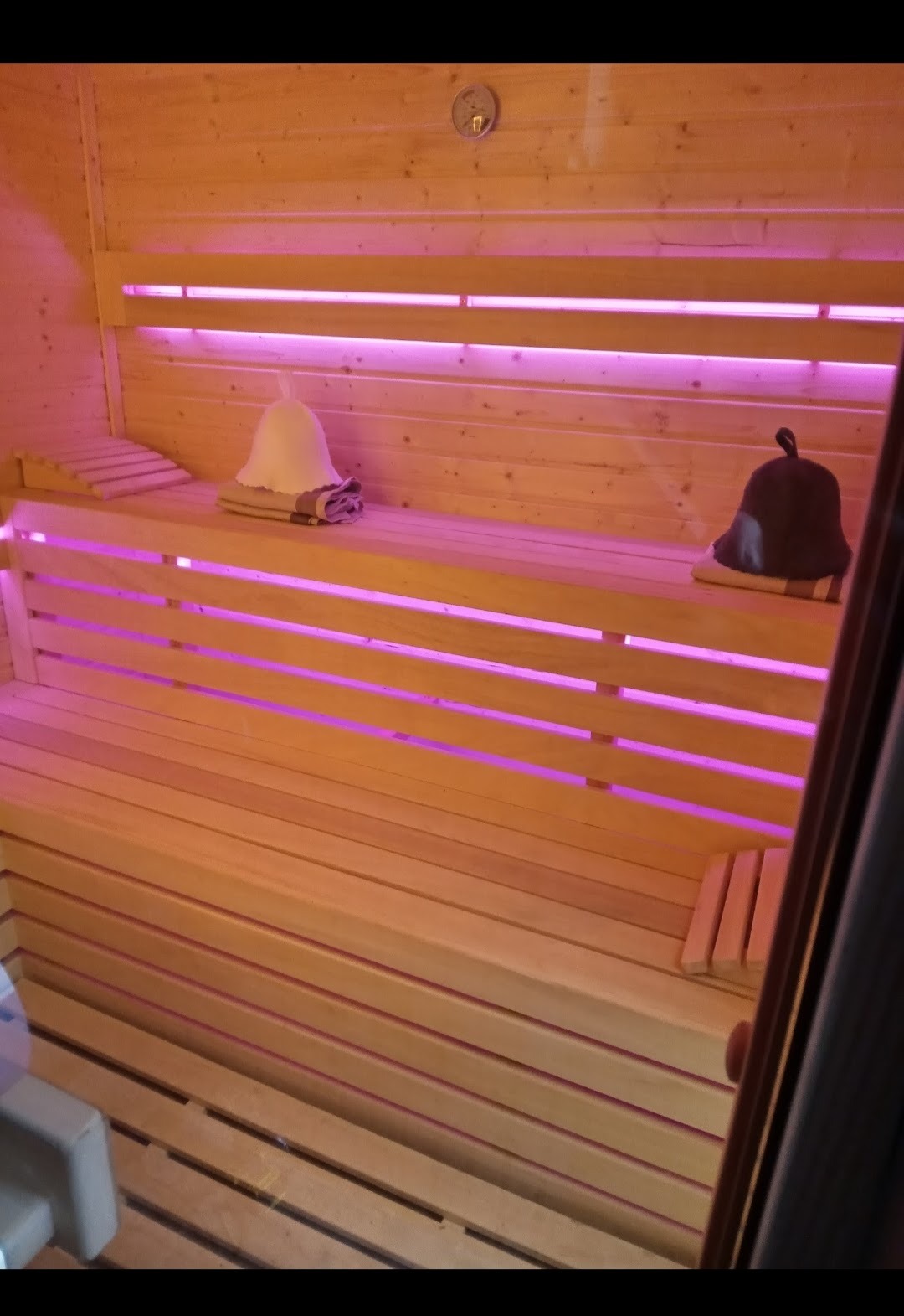 Wnętrze sauny z drewnianym wykończeniem, podświetleniem LED w kolorze fioletowym, na ławach leżą czapki i ręczniki.