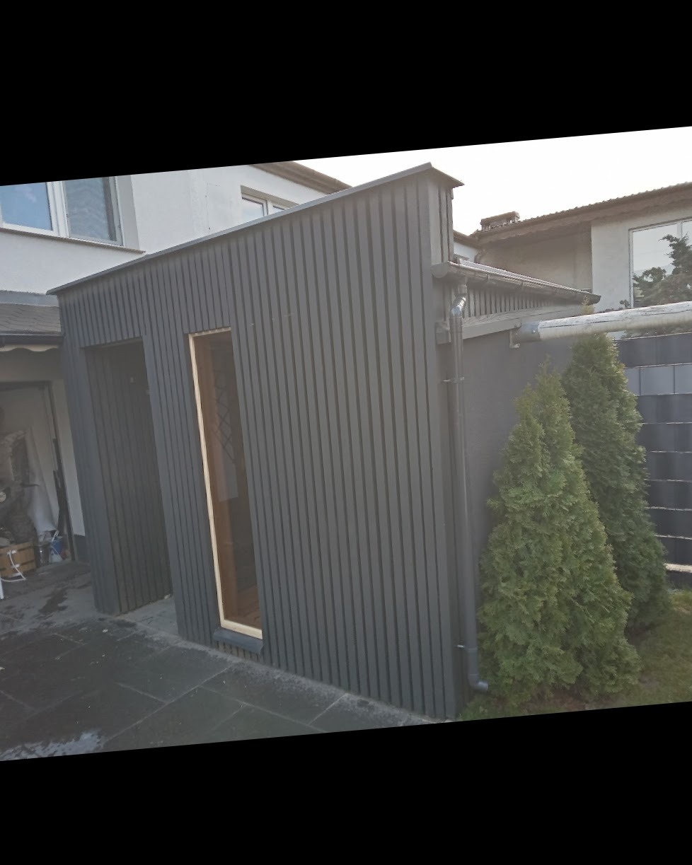 Szara konstrukcja z pionowych desek z oknem i drzwiami, prawdopodobnie szopa lub domek narzędziowy, przylegająca do domu, z dwiema tujami obok.