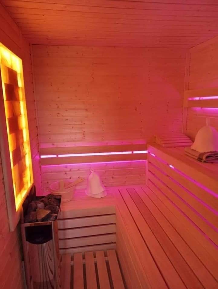 Wnętrze sauny wykończone jasnym drewnem, oświetlone nastrojowym, pomarańczowo-różowym światłem. Widoczne drewniane ławki, piec z kamieniami, drewniane wiadro z chochlą oraz filcowy kapelusz na głowę.