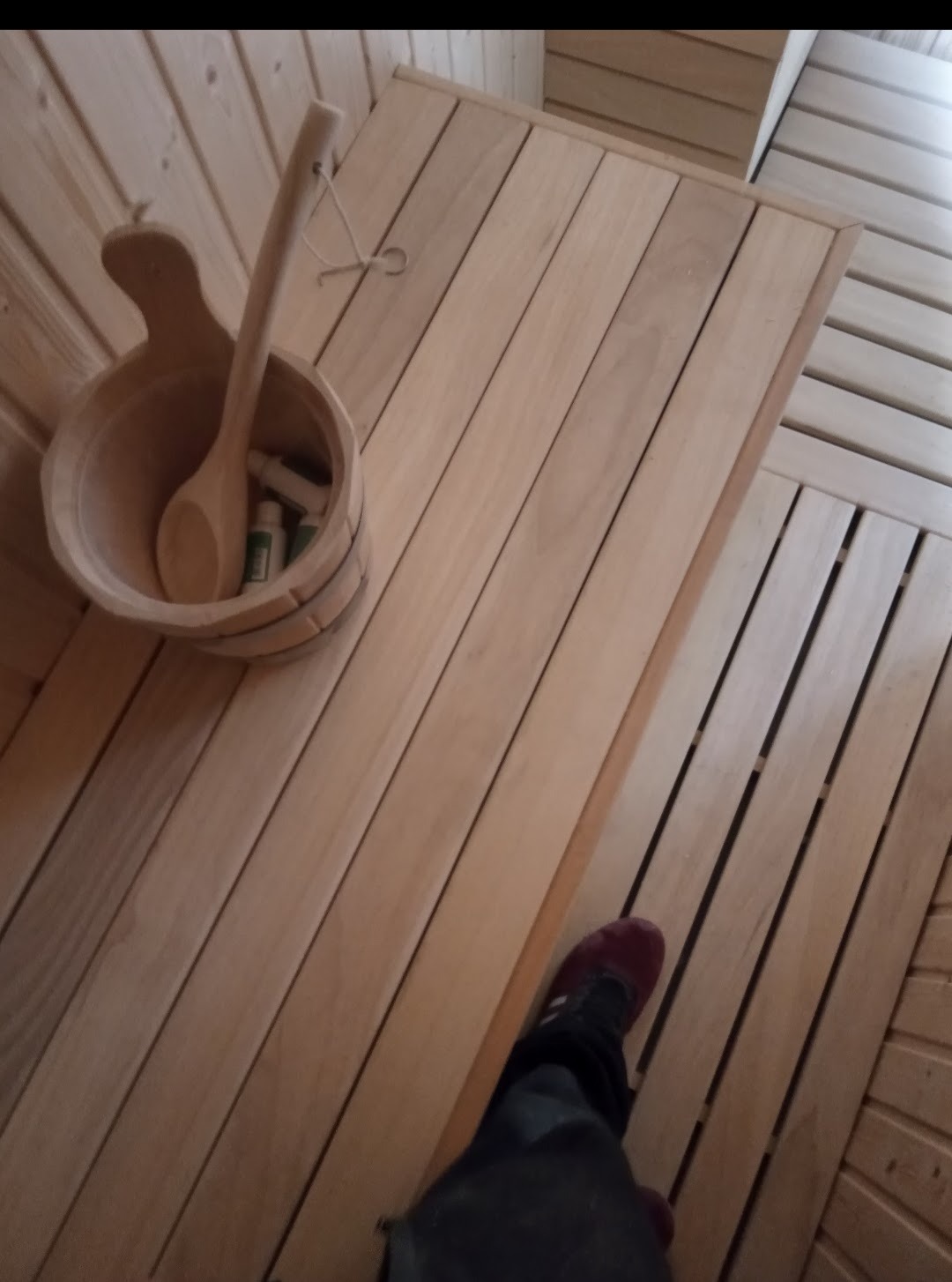 Wnętrze sauny z drewnianym wykończeniem, widoczny ceber z chochlą, butelki kosmetyków i fragment czyjejś nogi w ciemnych spodniach i bordowym bucie.