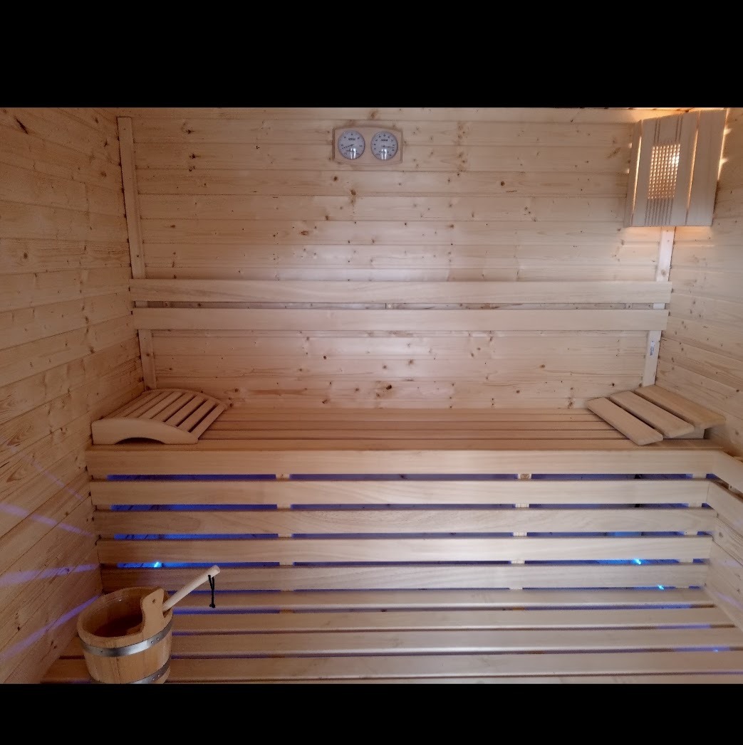 Wnętrze sauny wyłożone jasnym drewnem, z drewnianymi ławkami podświetlonymi na niebiesko, drewniany ceber z chochlą w rogu, dwa okrągłe termometry na ścianie.