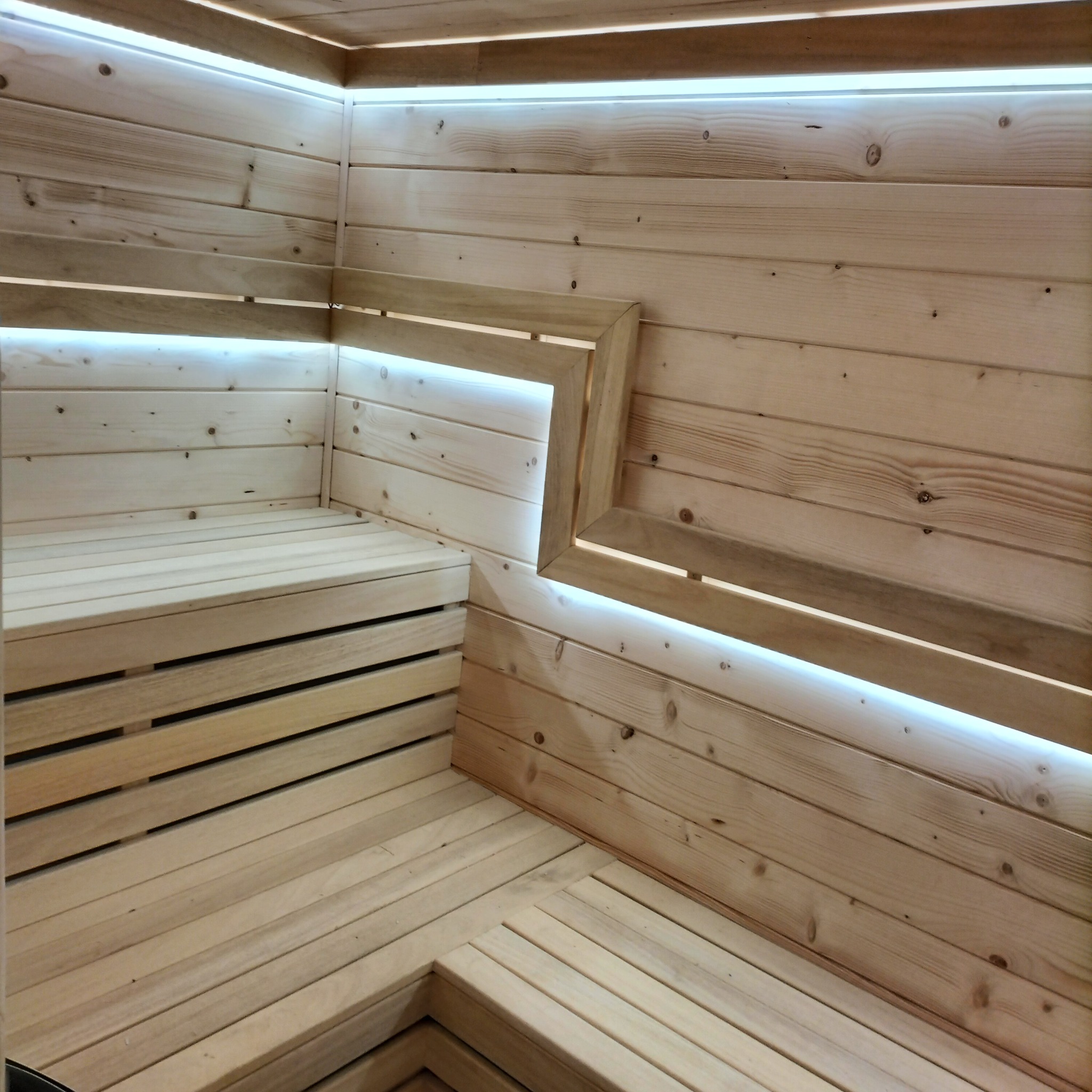 Wnętrze sauny wykończone jasnym drewnem, z podświetleniem LED wzdłuż górnej krawędzi i w narożnikach, ukazujące ławki i podłogę z desek.
