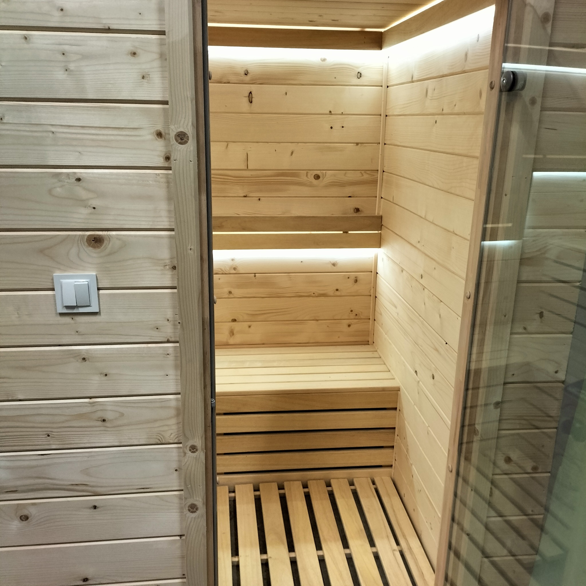 Wnętrze sauny z jasnego drewna z podświetleniem LED pod górną ławką, widoczne drewniane ławki i podłoga z listew, częściowo otwarte drzwi z szybą.