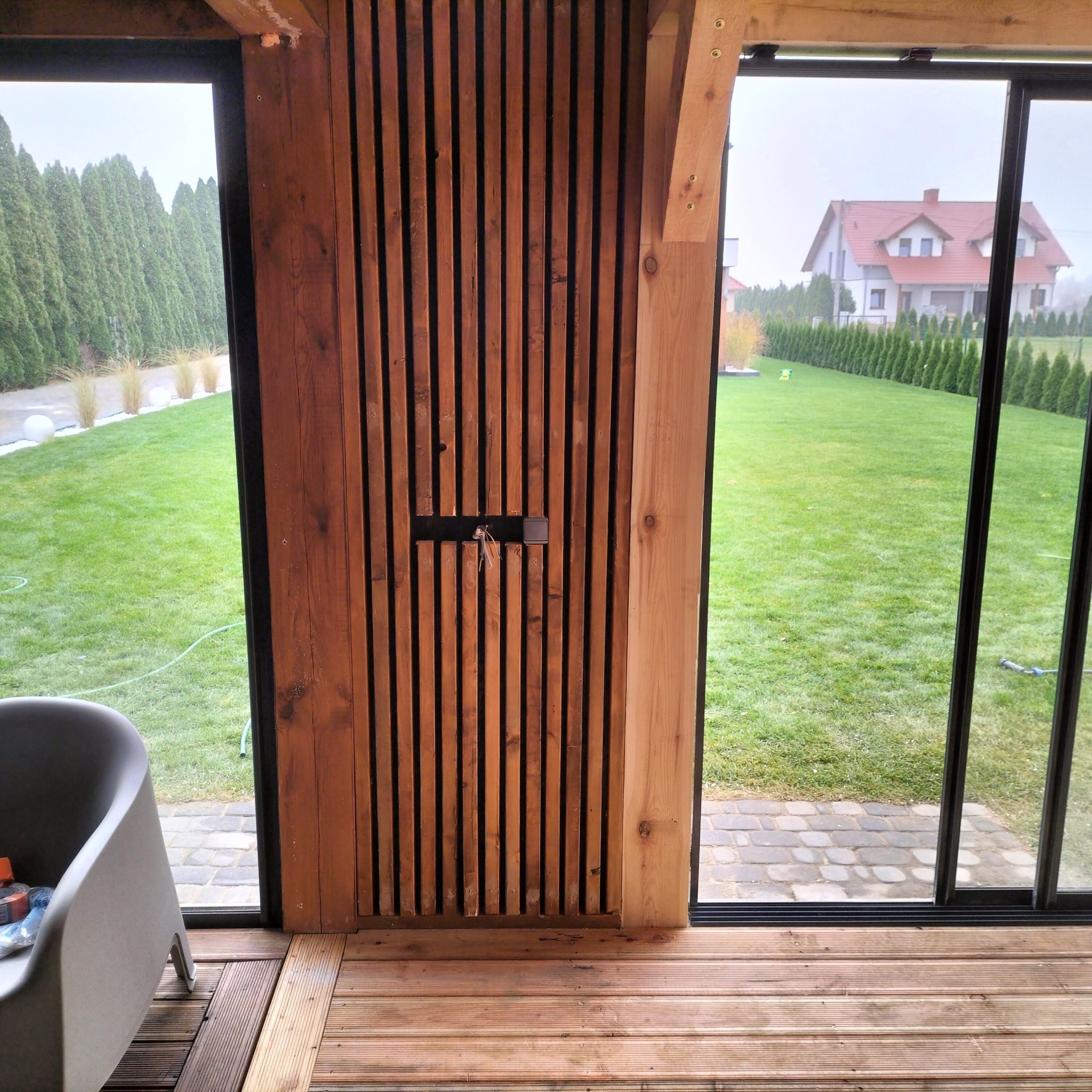 Nowoczesna weranda z drewnianą podłogą i ścianą z pionowych desek, widok na zadbany trawnik i dom w tle, szare krzesło w rogu kadru.