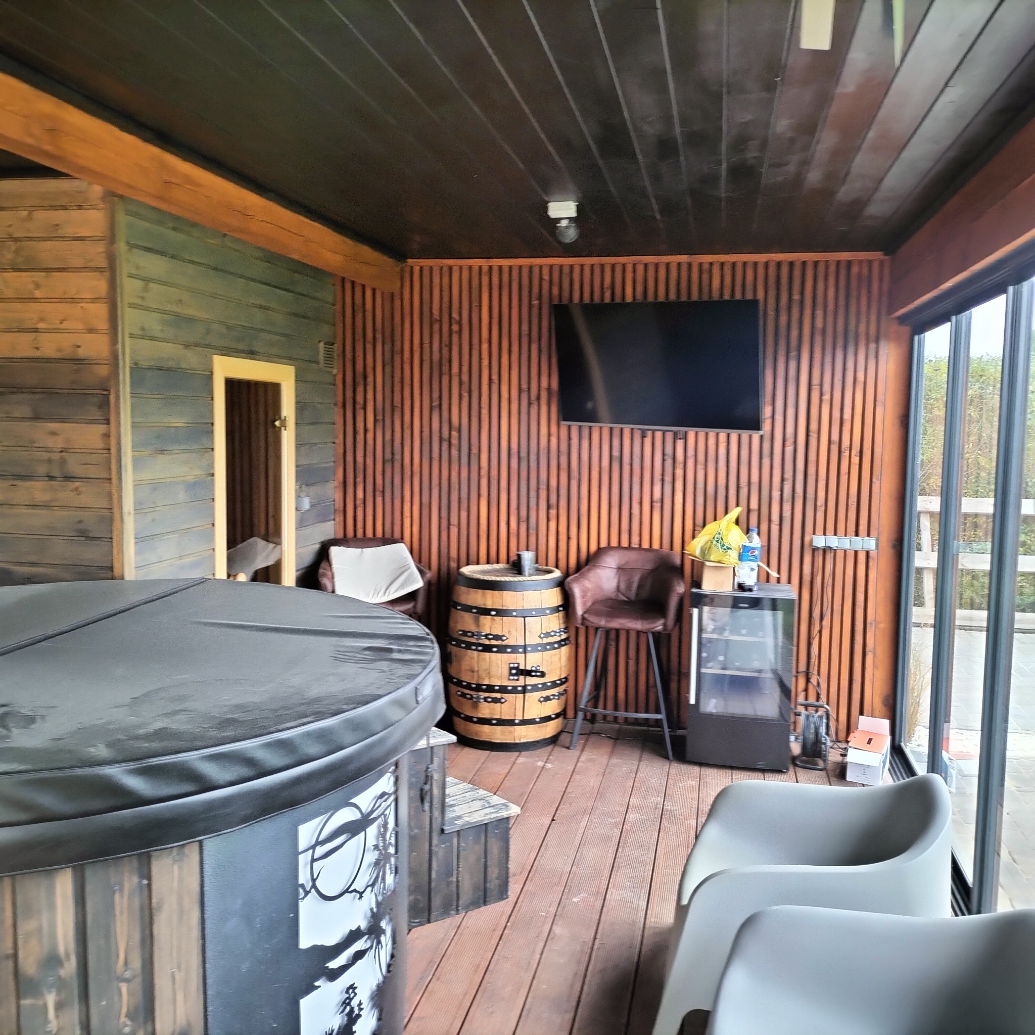 Wnętrze drewnianej sauny z jacuzzi, beczką pełniącą funkcję stolika, krzesłami barowymi i telewizorem na ścianie z drewnianych paneli.