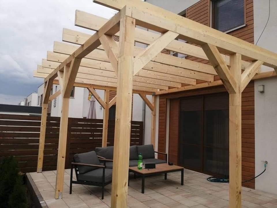 Surowa drewniana konstrukcja zadaszenia tarasu, z widocznymi belkami i słupami, na wybrukowanym patio z szarymi meblami ogrodowymi i niskim stolikiem kawowym, na tle domu z drewnianą elewacją...