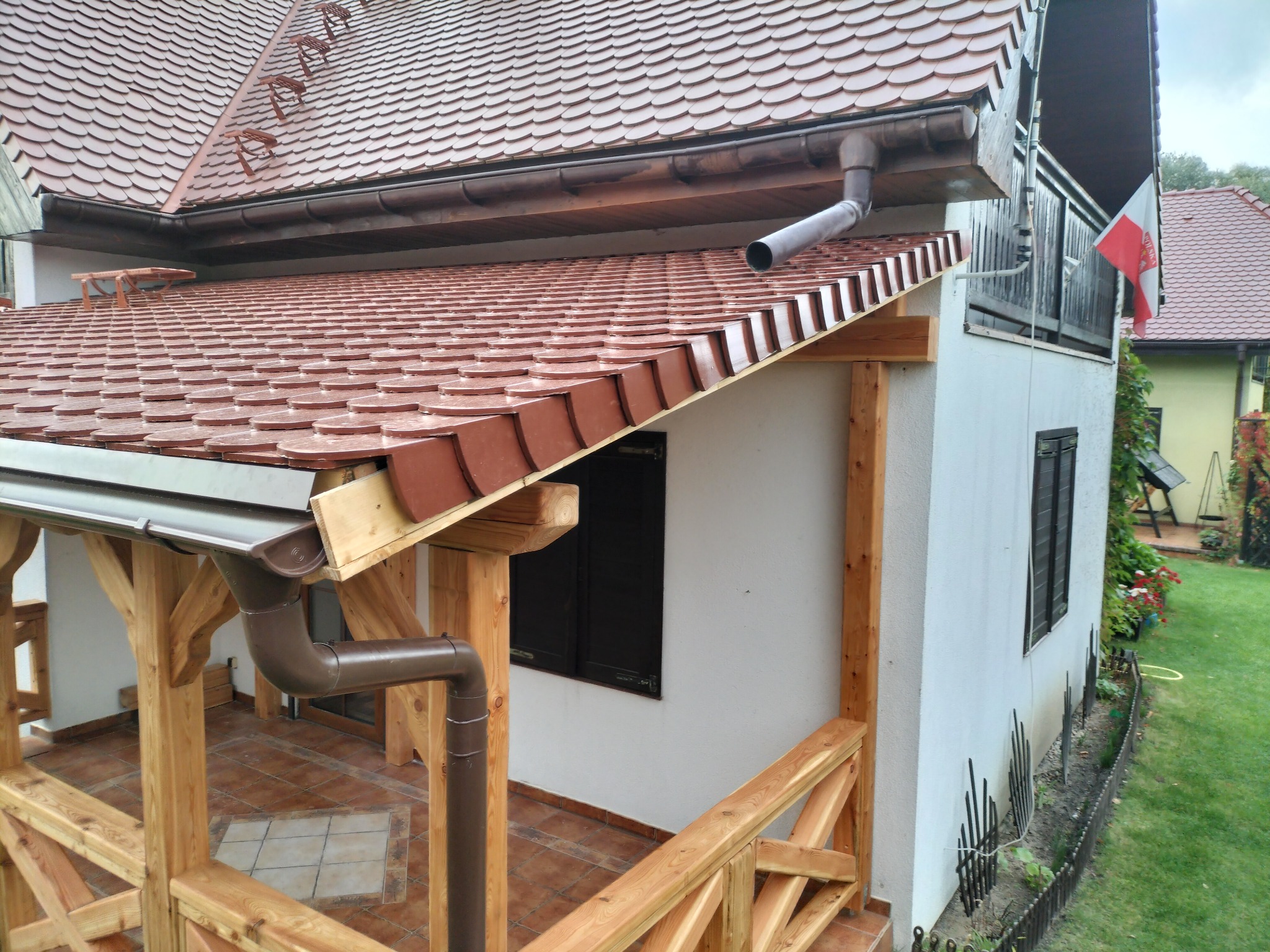 Zadaszony taras z drewnianą konstrukcją, brązowymi rynnami i ceramiczną podłogą, przylegający do białego budynku z oknami z okiennicami i polską flagą na balkonie.