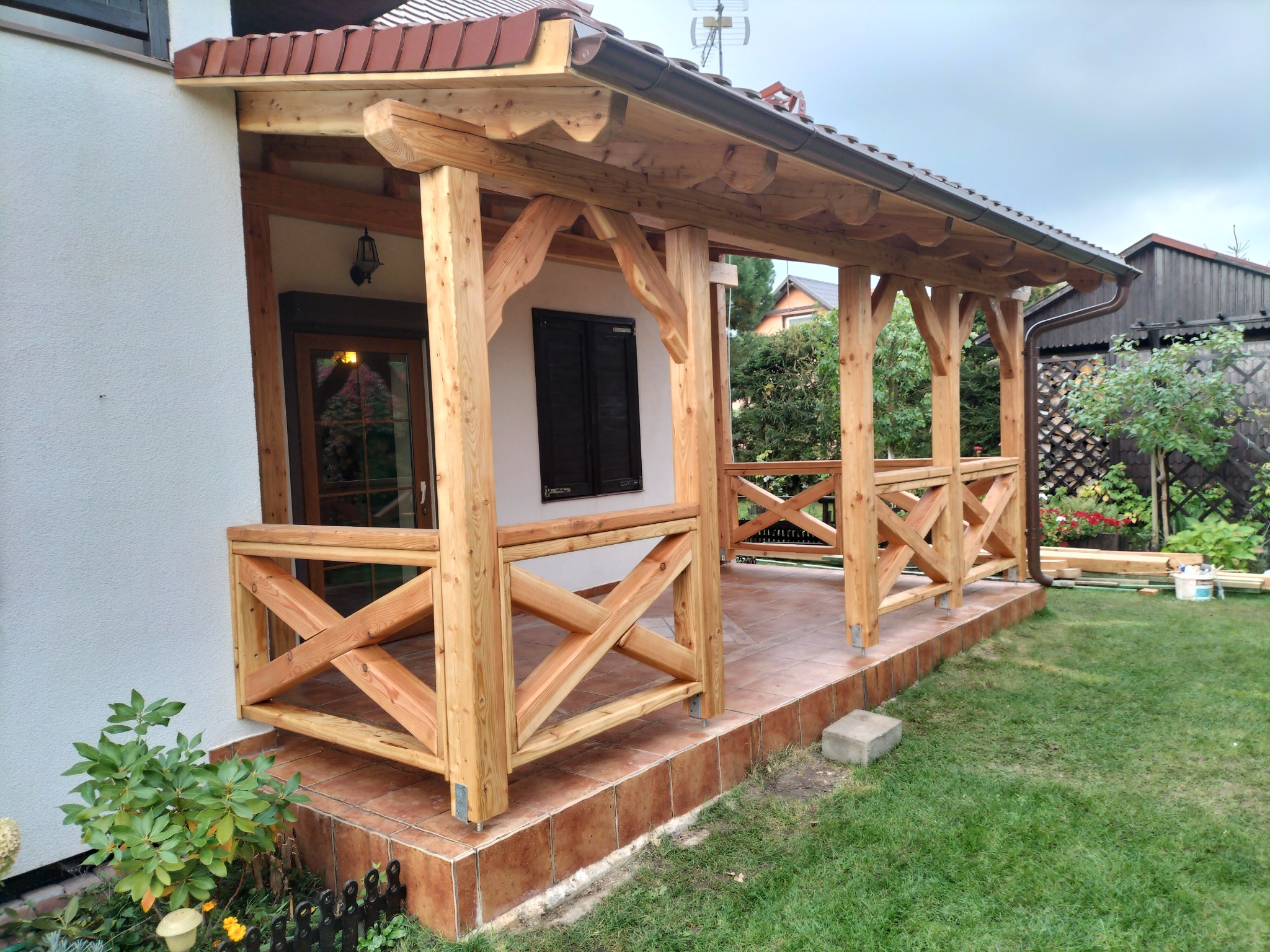 Zadaszony drewniany ganek z balustradą w kształcie krzyży, wykończony brązową dachówką, przylegający do białego budynku z brązowymi drzwiami i okiennicami, otoczony zielonym trawnikiem.